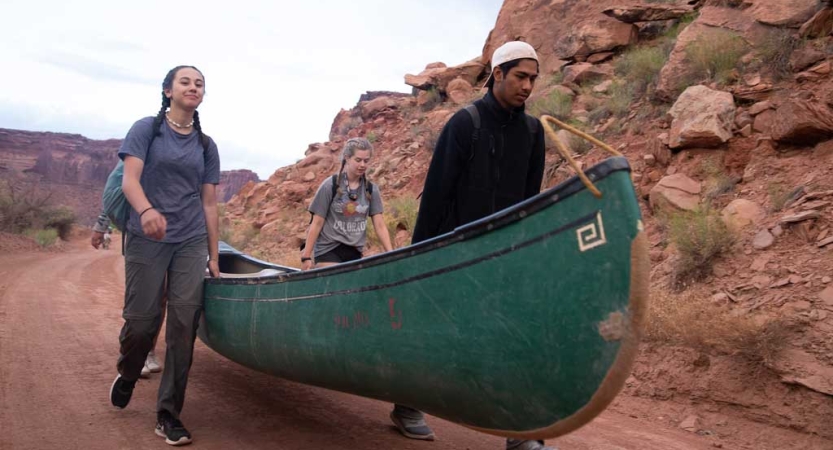 Three people carry a canoe along a dirt road